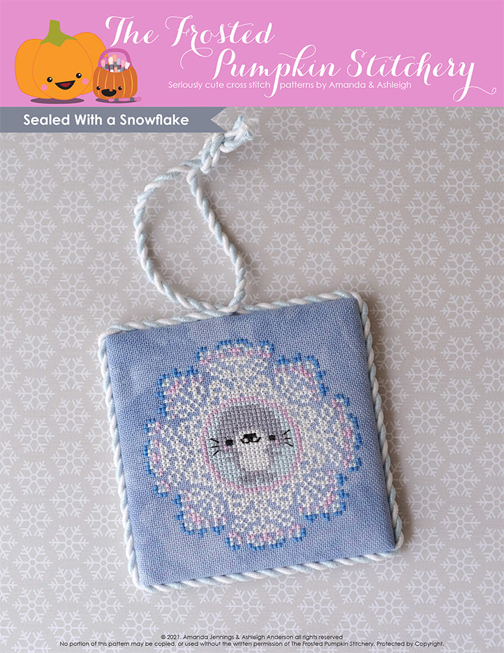 A little gray seal is stitched in the center of a snowflake with pinks and blues on gray fabric. It's finished as a square flat fold with white trim.
