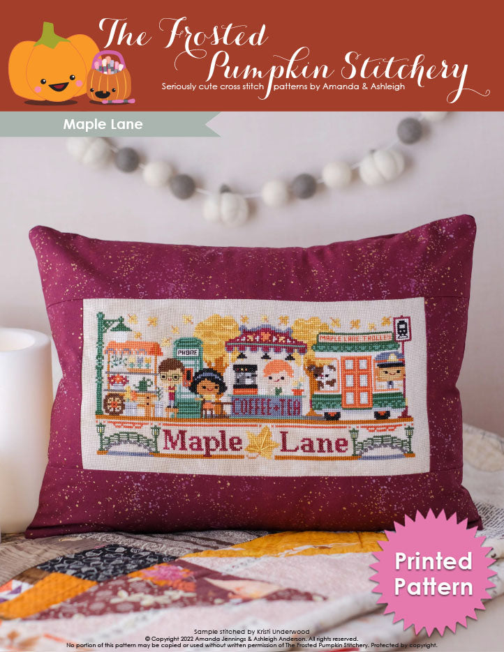 Maple Lane Cross Stitch Pattern. Cover image includes a barista working at the Coffee & Tea Bar, phone booth, a customer sitting at a table with her drink, the Maple Lane Trolley and a flower cart. They are surrounded by maple trees and floating maple leaves. This pattern is finished as a throw pillow.