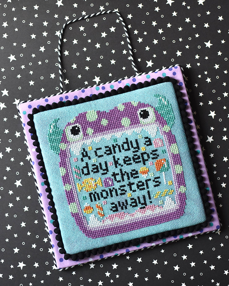 A flat lay of a cross stitch laying on black paper with white stars. The fabric is aqua and it is stitched with the phrase "a candy a day keeps the monsters away"