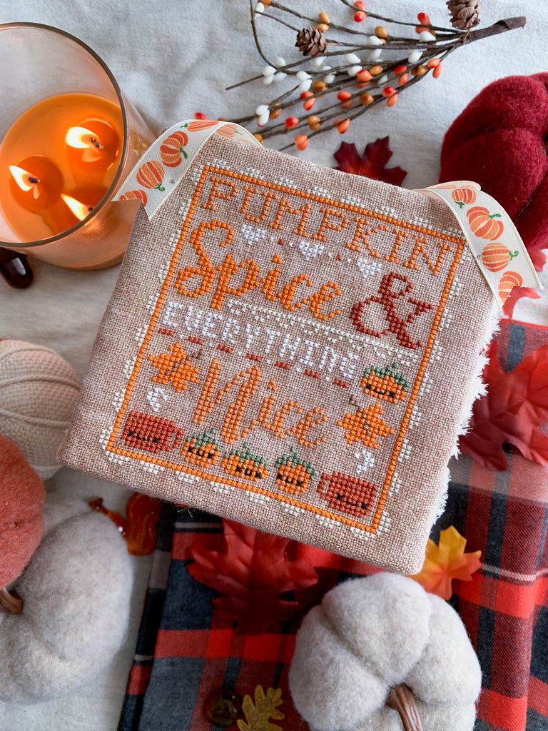 Image of Pumpkin Spice and Everything Nice Cross Stitch Pattern. Surrounded by plaid, pillows and felt pumpkins, along with a lit candle. 