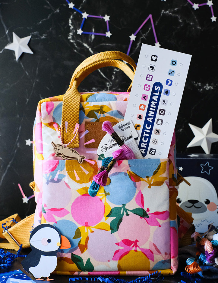 A backpack surrounded by embroidery floss skeins, blue crinkle paper and paper cut puffin  is pictured against a starry black background. A woodcut narwhal scissor fob is looped through the bag's handle.