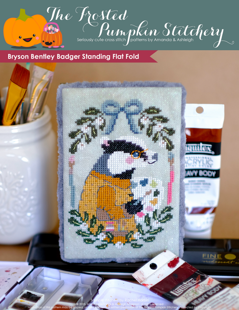 Cross-stitched flat fold with a european badger on a table with art and painting supplies.