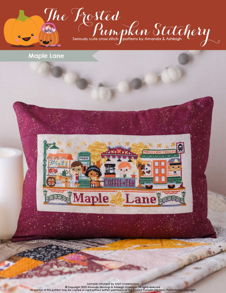 Maple Lane Cross Stitch Pattern. Cover image includes a barista working at the Coffee & Tea Bar, phone booth, a customer sitting at a table with her drink, the Maple Lane Trolley and a flower cart. They are surrounded by maple trees and floating maple leaves. This pattern is finished as a throw pillow.