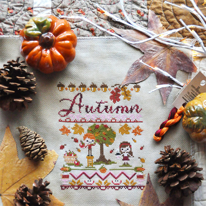 Flat lay image of Autumn Harvest Festival counted cross stitch pattern on a quilt with pine cones, leaves, a ceramic pumpkin.