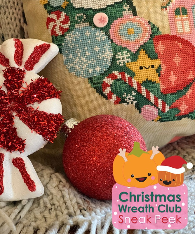 Christmas Wreath Club Sneak Peek. Two Christmas ornaments are balanced next to the corner of the pillow. The pillow is cross stitched with candy canes, a bow and ornaments.