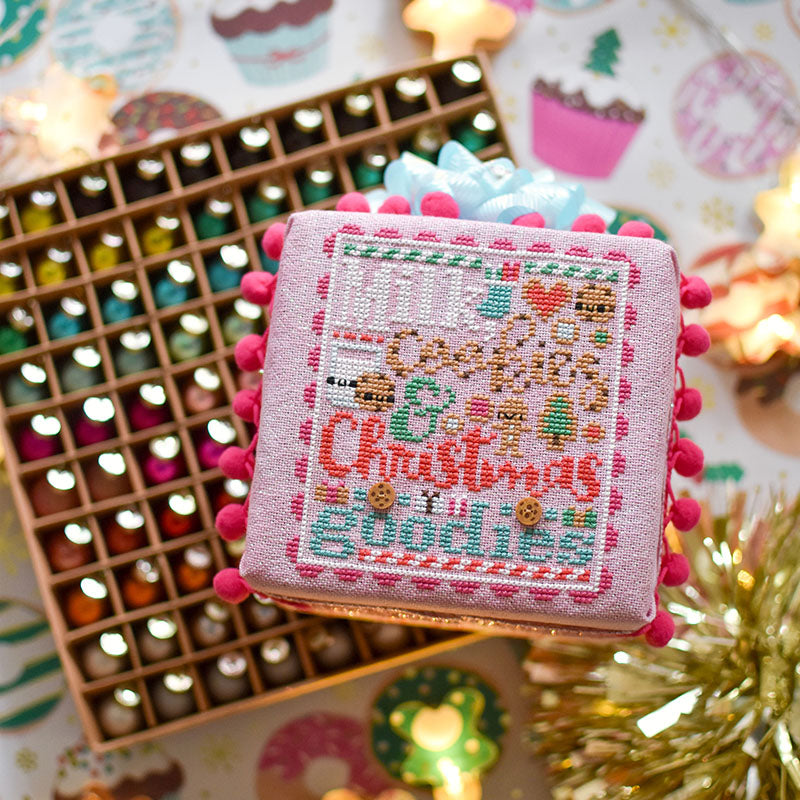Counted cross stitch pattern. Text reads Milk, Cookies and Christmas Goodies. A finished cross stitch pattern on a foam cube is laying on a box of ornaments.