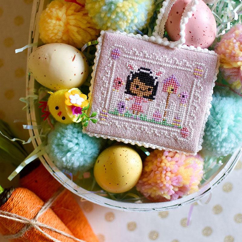 Easter Egg Hunt Counted Cross Stitch Pattern. A dark skinned girl with black hair is wearing bunny ears and Easter egg hunting with some small chicks. It's finished as a flat fold and there are pom poms in a basket.