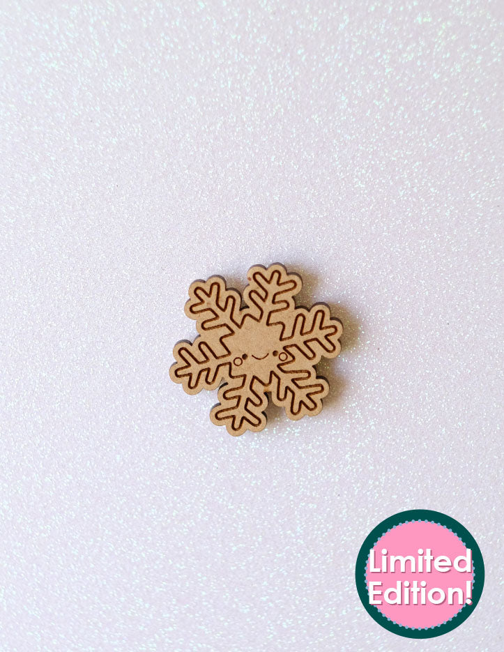 Flurry the Snowflake Needleminder. A wooden needleminder in the shape of a snowflake with a smile and round cheeks is sitting on a piece of sparkly white paper.