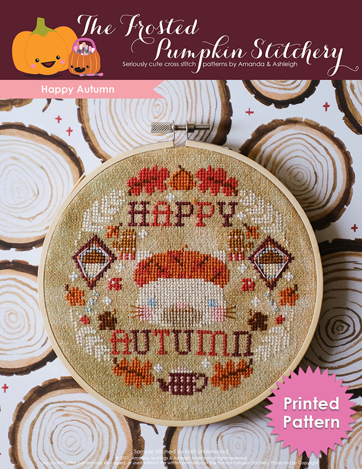An embroidery hoop with a cream colored cat wearing a beret surrounded by leaves, kites, mittens, wheat, a pumpkin and tea pot and the text "Happy Autumn." Printed Pattern.