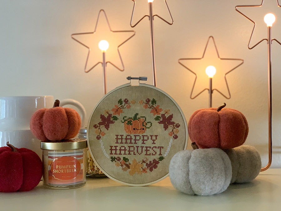 Happy Harvest cross stitch pattern. Design is a pumpkin with a face, surrounded by leaves and acorns. It's in an embroidery hoop surrounded by felt pumpkins, a candle and some glow-y star lights.