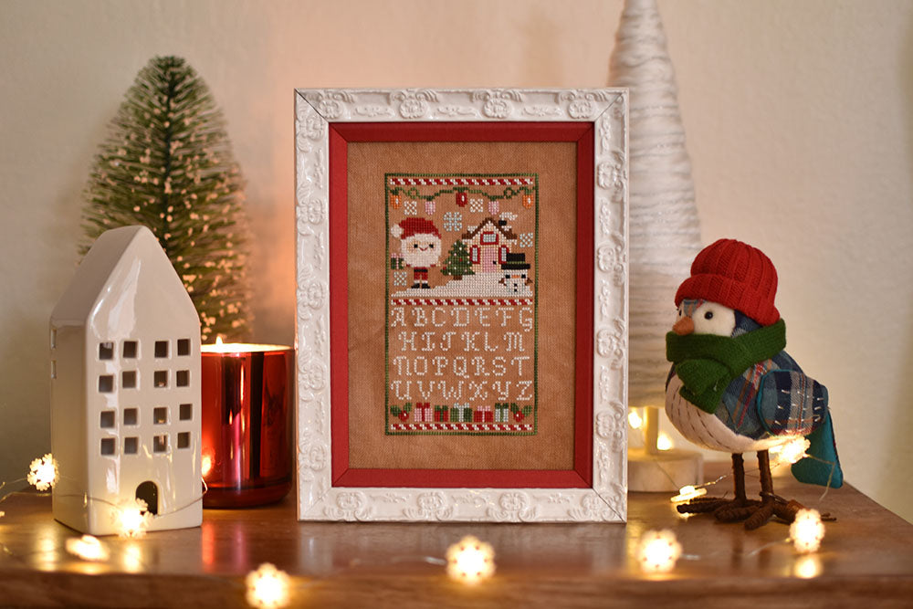 Holly Jolly Sampler counted cross stitch pattern. Christmas pattern with santa, a snowman and a little house above the alphabet. Surrounded by a ceramic house, a fake bird wearing a hat and a bristle tree.