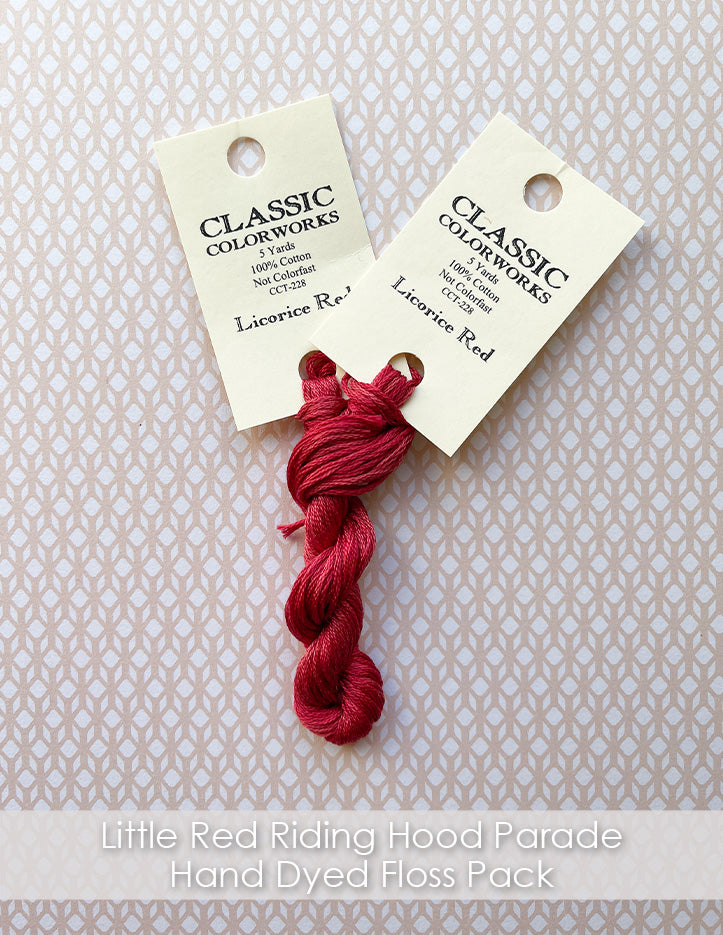 Two skeins of red hand-dyed embroidery floss are twisted together with their cream color tags reading "Classic Colorworks Licorice Red" on peach patterned paper.