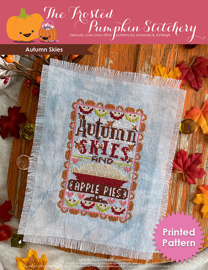 Autumn Skies counted cross stitch pattern. Text reads Autumn Skies and Apple Pies. Image of blue linen with the pattern cross stitched onto it, a big apple pie in the center, pie slices on the borders. Printed Pattern.