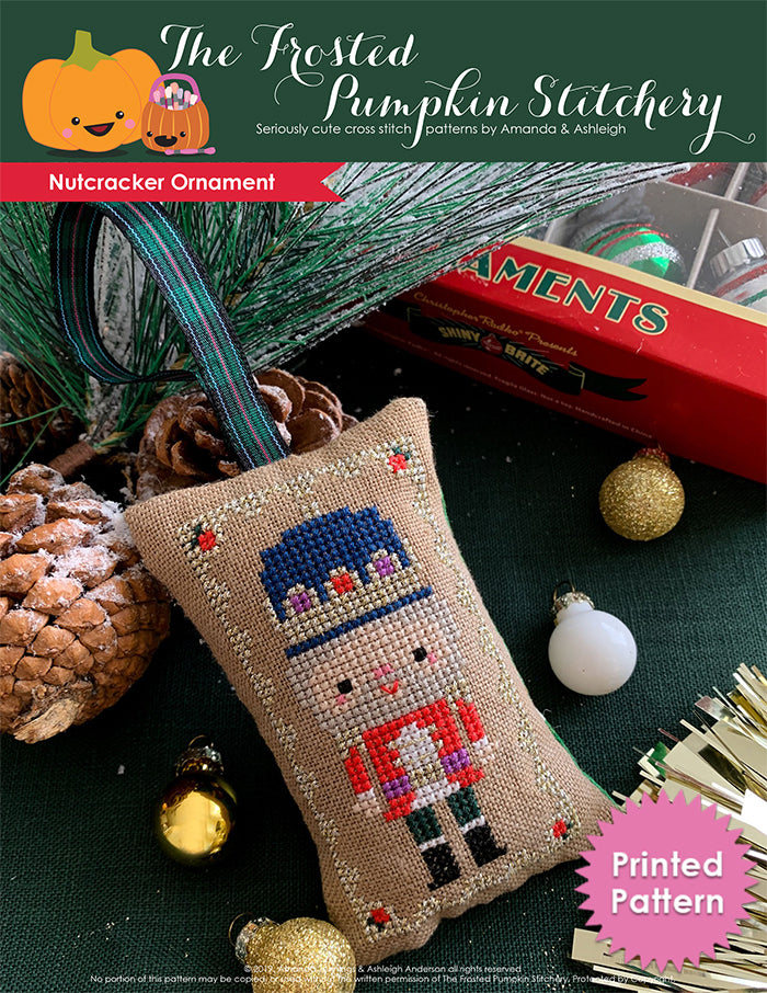 Nutcracker Ornament counted cross stitch pattern. A white skinned nutcracker with gray hair in a red uniform with a gold border. Laying on a piece of green fabric surrounded by pinecones and ornaments. Printed Pattern.