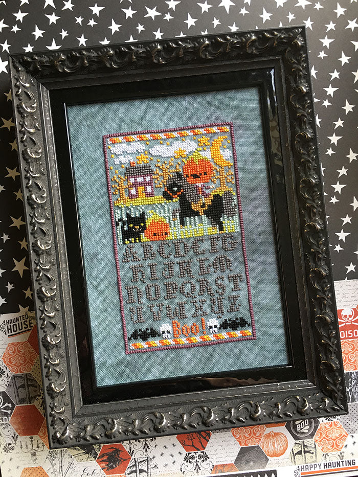 Sleepy Hollow Sampler counted cross stitch pattern. The headless horsemen is riding in the village at night. Alphabet below in a Halloween inspired font. Framed in a gothic thick black frame.