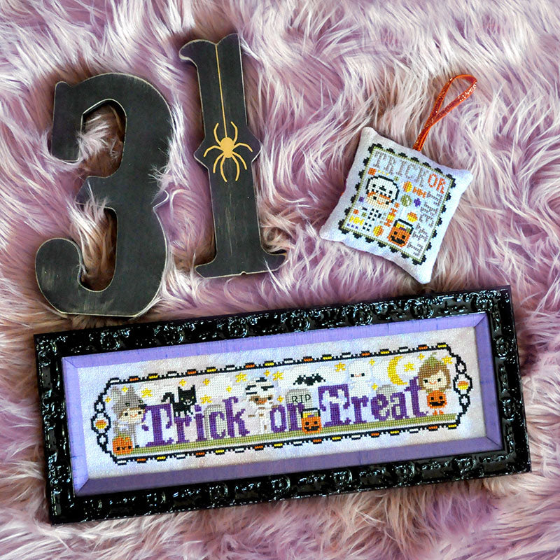 Trick or Treat Costume Club counted cross stitch pattern. Three trick-or-treaters dressed as a wolf, a mummy and a pumpkin. Text says "Trick or Treat". Wooden numbers 31 for Halloween are laying next to the pattern.