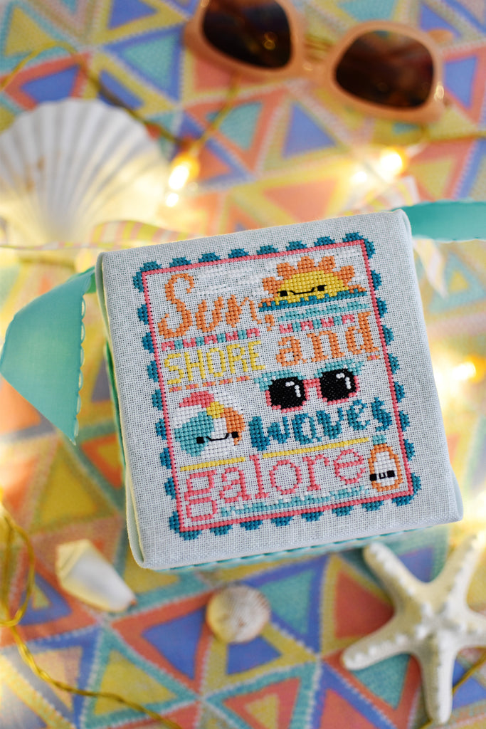 Waves Galore Counted Cross Stitch Pattern. Image of a kawaii sun, beach ball, sunglasses and sunscreen. Tropical colors including aqua, bright pink and oranges. Text in the chart reads Sun, Shore and Waves Galore. Background of the photo are sunglasses and seashells.