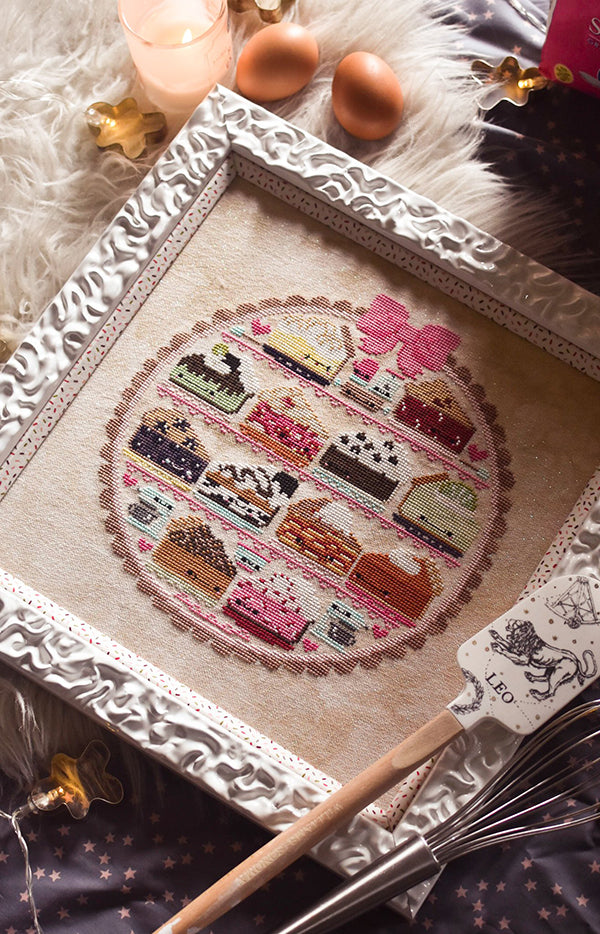 Sweet as Pie counted cross stitch pattern. Twelve kawaii pies cross stitch pattern in a white frame surrounded by baking supplies. Flay lay on a fake fur rug with eggs.