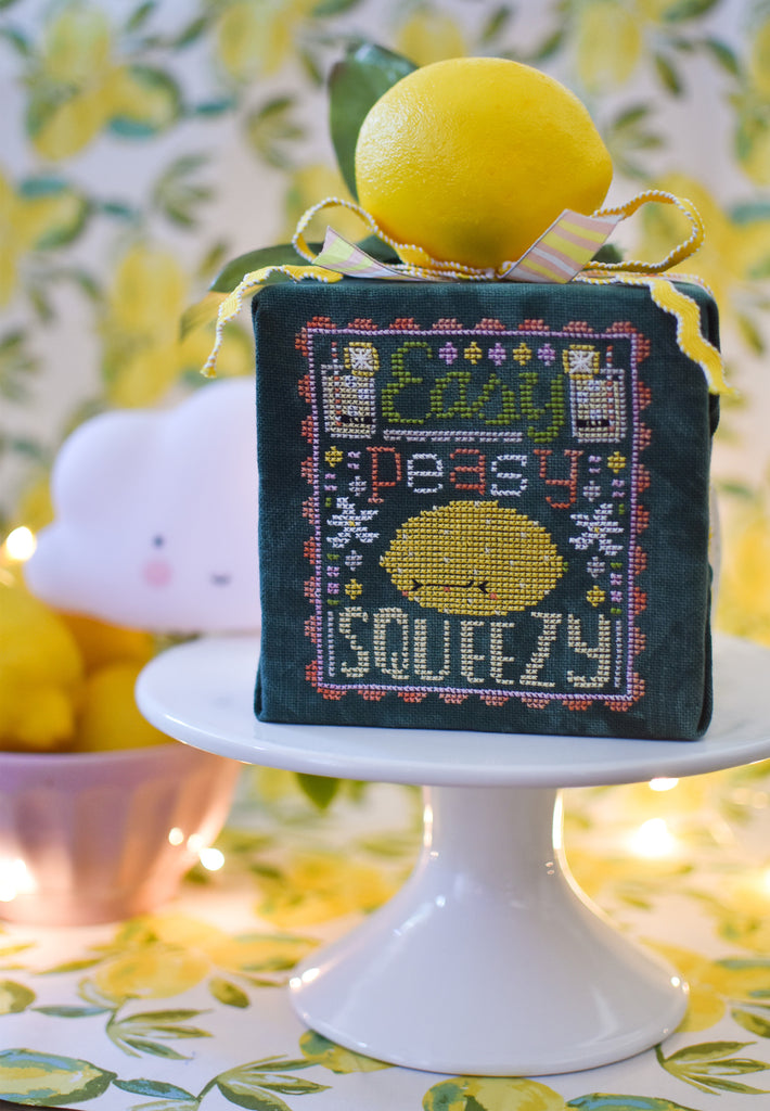 Easy Peasy Counted Cross Stitch Pattern. Image of a sour faced lemon stitched on green fabric with the text Easy Peasy above the lemon and below the word Squeezy. A background of lemons.