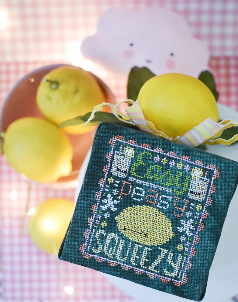 Easy Peasy Counted Cross Stitch Pattern. Image of a sour faced lemon stitched on green fabric with the text Easy Peasy above the lemon and below the word Squeezy. Image background is pink gingham, a little cloud and a bowl of lemons.