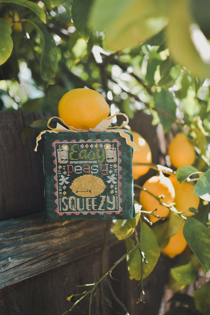 Easy Peasy Counted Cross Stitch Pattern. Image of a sour faced lemon stitched on green fabric with the text Easy Peasy above the lemon and below the word Squeezy. Photo taken on a summer morning with glow-y light and a lemon tree in the background.