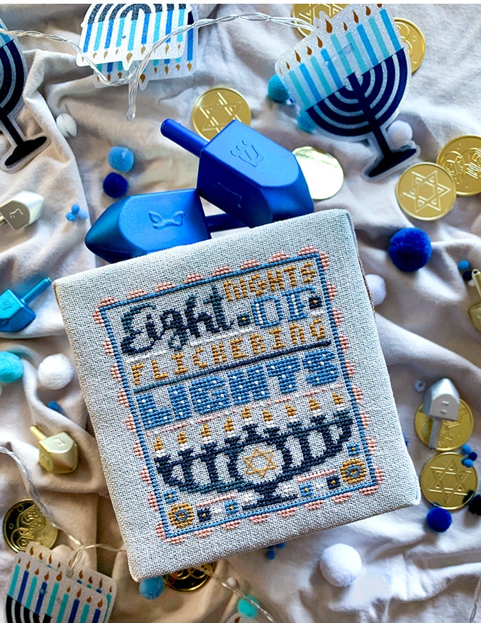 Eight Nights Counted Cross Stitch Pattern. Text reads "Eight nights of flickering lights" with a menorah. Background is gelt, dreidels and menorah lights.