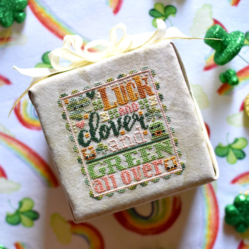 Luck and Clover counted cross stitch pattern. St. Patrick's Day inspired with pinks, golds, dark and light green. Text reads "Luck and Clover and Green All Over". Finished project is laying on a rainbow background.
