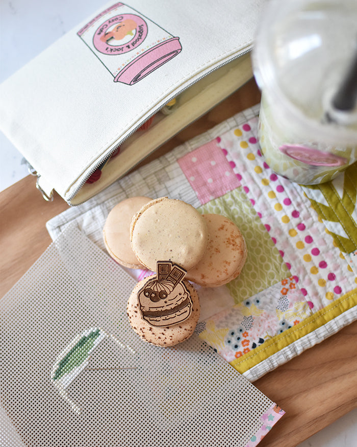 A matcha cup is being stitched on perforated paper next to macarons on a quilted patchwork mat in greens and pinks.