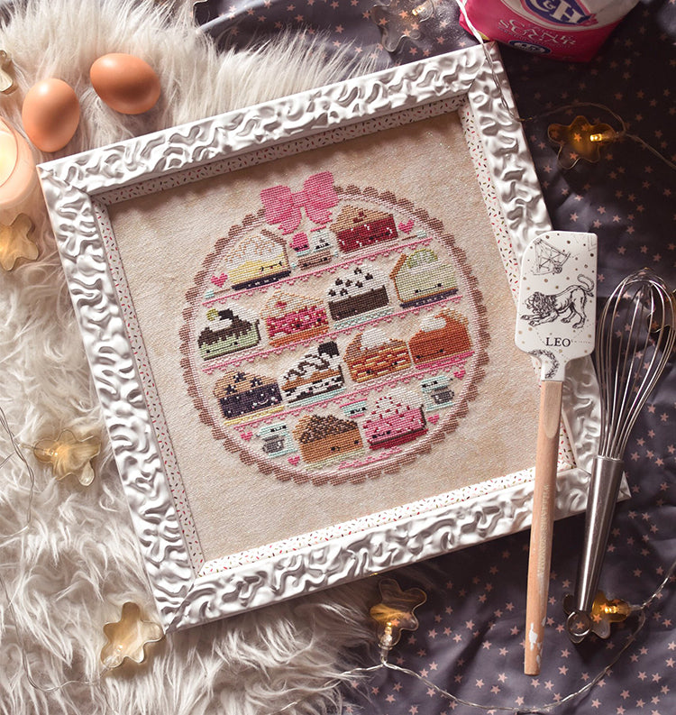 Sweet as Pie counted cross stitch pattern. Twelve kawaii pies cross stitch pattern in a white frame surrounded by baking supplies. Flat lay on a gray background with a spatula that reads "Leo".